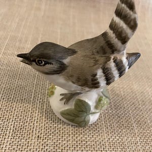 May include: A ceramic bird ornament with a detailed design. The bird has a grey head and back, with a striped tail. It is perched on a white base with green leaves and berries. The ornament is a decorative item.