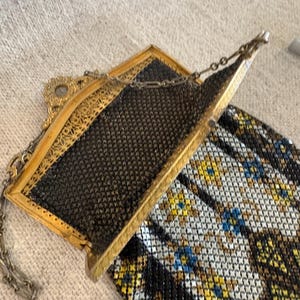 May include: An antique gold-toned metal mesh purse with a black mesh interior and a decorative frame. The purse has a chain handle and a colorful patterned bottom with blue, yellow, and white accents. A vintage accessory.