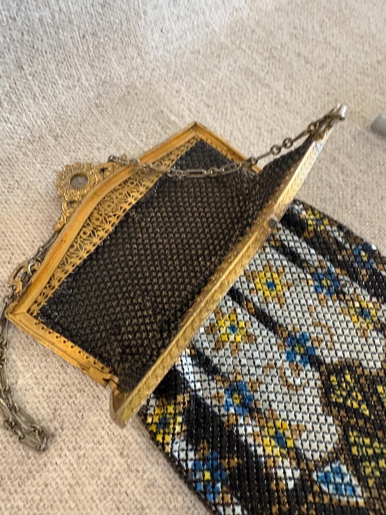 May include: An antique gold-toned metal mesh purse with a black mesh interior and a decorative frame. The purse has a chain handle and a colorful patterned bottom with blue, yellow, and white accents. A vintage accessory.