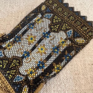 May include: An antique metal mesh purse with a decorative floral pattern in white, yellow, and blue. The purse has a black border and a gold-colored filigree frame. The purse is on a neutral-colored fabric.