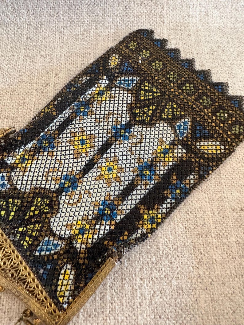 May include: An antique metal mesh purse with a decorative floral pattern in white, yellow, and blue. The purse has a black border and a gold-colored filigree frame. The purse is on a neutral-colored fabric.