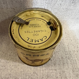 May include: Vintage Camel cigarettes tin. The tin is cylindrical with a yellow and brown color scheme. The lid features the text "100 Cigarettes" and "Camel" in a circular design. The side of the tin displays a camel silhouette.