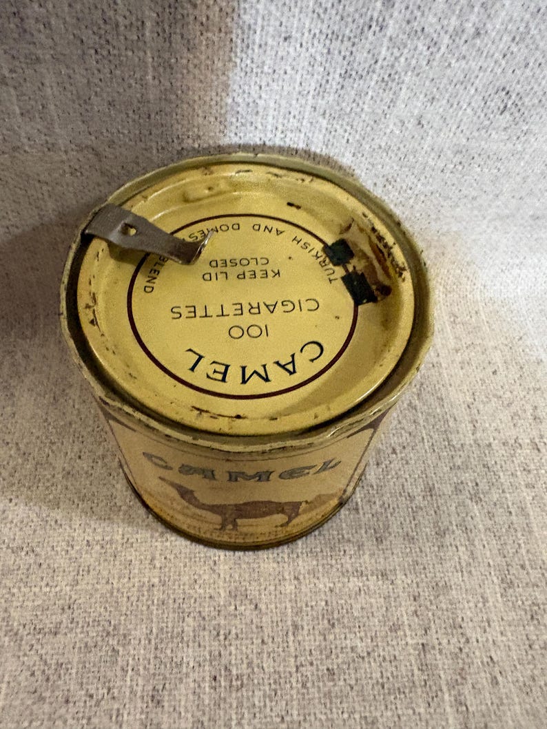 May include: Vintage Camel cigarettes tin. The tin is cylindrical with a yellow and brown color scheme. The lid features the text "100 Cigarettes" and "Camel" in a circular design. The side of the tin displays a camel silhouette.