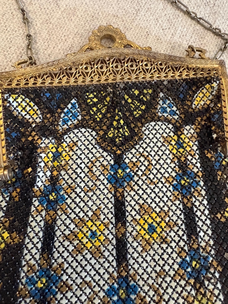 May include: An antique metal mesh handbag with a gold-toned frame and chain. The mesh is black, white, yellow, and blue, with a floral pattern. The frame has an ornate, filigree design.