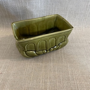 May include: A rectangular, olive green ceramic planter with a glossy finish. The planter features a decorative, repeating arched design along the base. The interior is dark, suggesting it is empty. The planter is suitable for small plants or succulents.