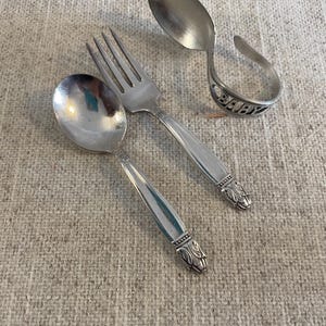 Pode incluir: Um conjunto de talheres de prata vintage para bebé, incluindo uma colher, um garfo e uma colher com uma pega curva. A pega curva da colher tem a palavra "BABY" gravada. Os talheres estão sobre uma superfície texturizada de cor neutra.