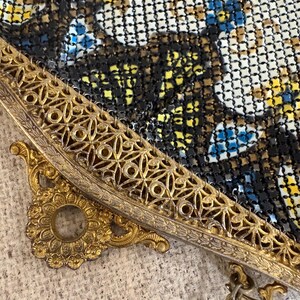 May include: Close-up of a vintage metal mesh purse. The purse features a gold-toned filigree frame and clasp. The mesh is a mix of black, white, yellow, and blue, creating a floral pattern. A delicate chain is attached.
