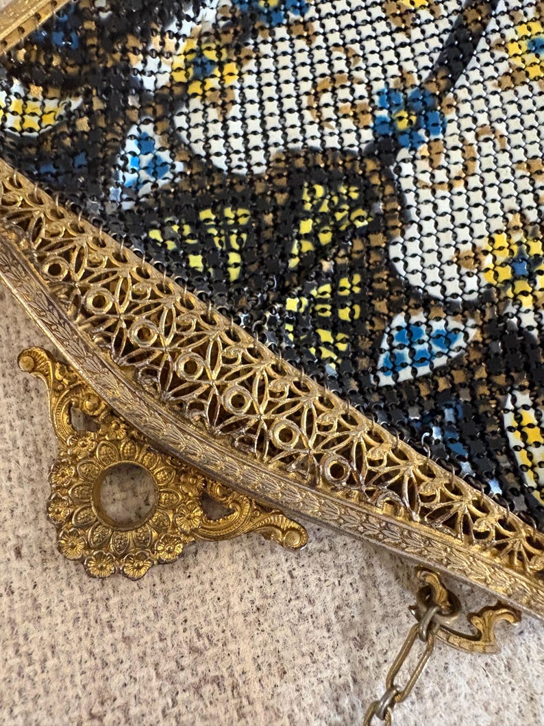 May include: Close-up of a vintage metal mesh purse. The purse features a gold-toned filigree frame and clasp. The mesh is a mix of black, white, yellow, and blue, creating a floral pattern. A delicate chain is attached.