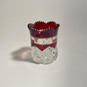 Op de afbeelding: Een kleine, heldere glazen vaas met een rode en gouden rand en een geslepen glaspatroon. De vaas is ongeveer 10 cm hoog.