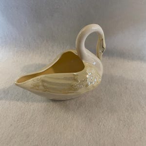 May include: A ceramic swan-shaped creamer or gravy boat. The swan is primarily off-white with a pale yellow interior and subtle iridescent sheen. The swan's neck curves gracefully, and the beak is detailed with black accents. The design is a classic, decorative piece.