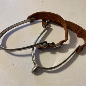 Könnte beinhalten: Ein Paar silberne Sporen mit braunen Lederriemen. Die Sporen haben ein spitzes Ende und eine kleine Metallschlaufe oben.
