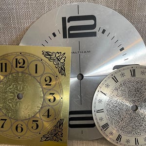 May include: Three clock faces in a close-up shot. One is gold with black numerals and floral corner designs. Another is silver with black numerals and the word "WALTHAM". The third is silver with black Roman numerals.