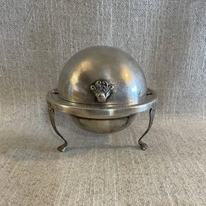Puede incluir: Una mantequera plateada con tapa abovedada y asa ornamentada. El plato se apoya sobre tres patas curvas. El diseño es vintage y elegante, adecuado para servir mantequilla en la mesa.