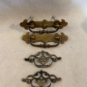 May include: Four antique brass drawer pulls. Two pulls have a rectangular backplate with a curved handle. The other two pulls have an ornate, openwork design. All pulls have a vintage aesthetic.