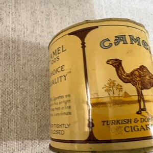 May include: A vintage Camel Turkish & Domestic Cigars tin. The tin is a light tan color with the word "Camel" in a teal font. The tin features an illustration of a camel, palm trees, and pyramids.
