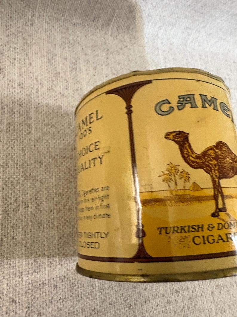 May include: A vintage Camel Turkish & Domestic Cigars tin. The tin is a light tan color with the word "Camel" in a teal font. The tin features an illustration of a camel, palm trees, and pyramids.