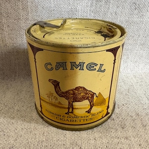 May include: Vintage Camel cigarette tin with a yellow and brown color scheme. The tin features the word "CAMEL" in large letters, a camel illustration, and text that reads "TURKISH & DOMESTIC BLEND CIGARETTES". The lid has a hinged closure.