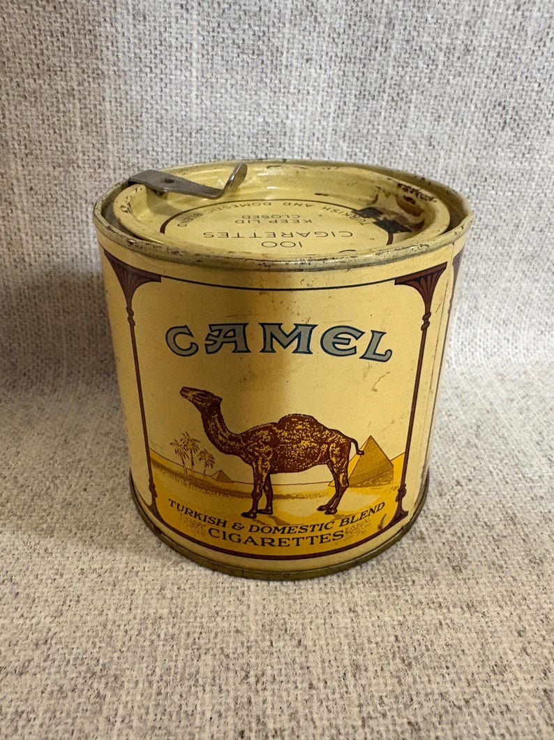 May include: Vintage Camel cigarette tin with a yellow and brown color scheme. The tin features the word "CAMEL" in large letters, a camel illustration, and text that reads "TURKISH & DOMESTIC BLEND CIGARETTES". The lid has a hinged closure.