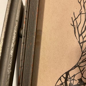May include: A black and white silhouette of a tree with branches extending out in a variety of directions. The image is framed in a dark wood frame with a red accent.