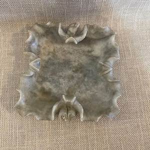 May include: A gray, leaf-shaped dish with carved bat and leaf designs. The dish has a scalloped edge and is made of a stone-like material. The dish is displayed on a neutral-colored, textured surface.