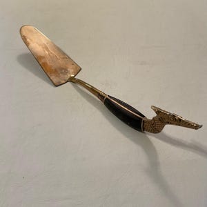 May include: A gold-toned cake server with a black handle and a dragon-shaped end. The server has a slightly curved blade.