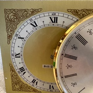May include: A vintage clock face with Roman numerals and a gold-tone bezel. The clock face is silver with black hour markers. A gold-colored paper with a clock face design is behind the clock. The paper has the text "Seth Thomas".
