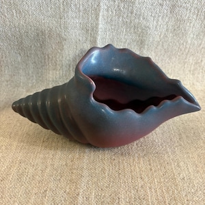 Puede incluir: Un cuenco decorativo de cerámica con forma de concha. El cuenco tiene un degradado de colores, con un exterior azul-verdoso y un interior rojizo. La concha tiene un diseño texturizado y acanalado en el exterior.