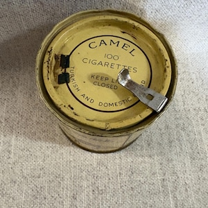 May include: Vintage Camel 100 Cigarettes tin. The tin is yellowed with age and has a metal opener attached. The text on the lid reads "Camel 100 Cigarettes" and "Keep Lid Closed". The tin is cylindrical.