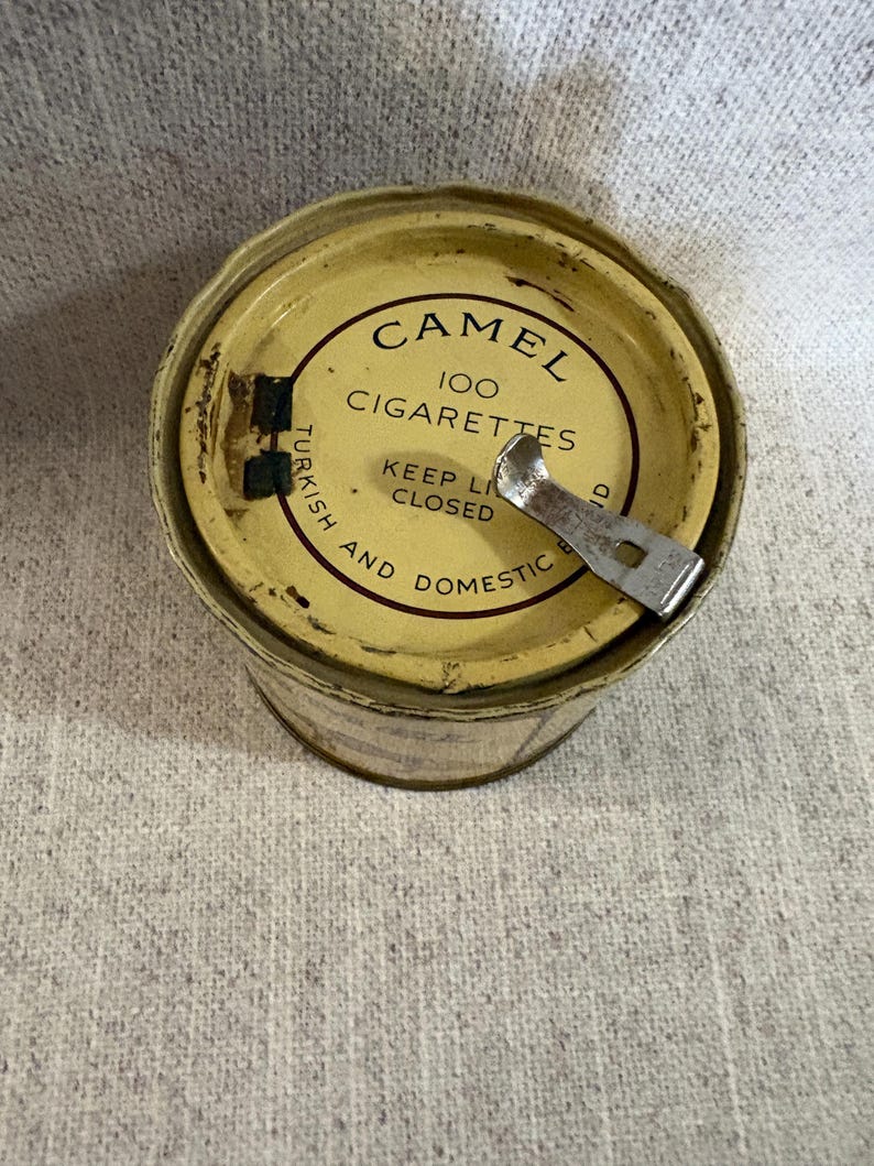 May include: Vintage Camel 100 Cigarettes tin. The tin is yellowed with age and has a metal opener attached. The text on the lid reads "Camel 100 Cigarettes" and "Keep Lid Closed". The tin is cylindrical.