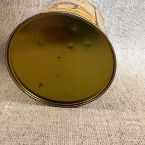 May include: An antique, cylindrical tin can with a gold-colored base and a slightly curved, beige-colored body. The body features a brown illustration and text. The can is resting on a textured, light gray surface.