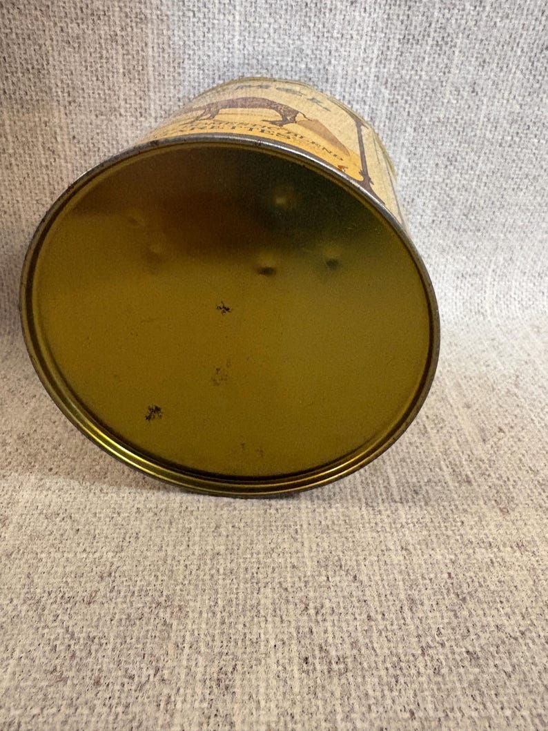May include: An antique, cylindrical tin can with a gold-colored base and a slightly curved, beige-colored body. The body features a brown illustration and text. The can is resting on a textured, light gray surface.