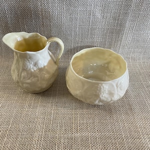 May include: Cream-colored ceramic creamer and sugar bowl set. The creamer has a handle and a spout. Both pieces have a textured, embossed design. The interior of both pieces has a light yellow hue. The set is on a neutral background.