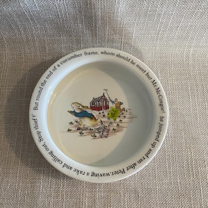 Może przedstawiać: Biały ceramiczny talerz z kolorową ilustracją królika uciekającego przed mężczyzną z grabiami. Talerz ma tekst wokół krawędzi, w tym "Stop thief!" i "Peter Rabbit".