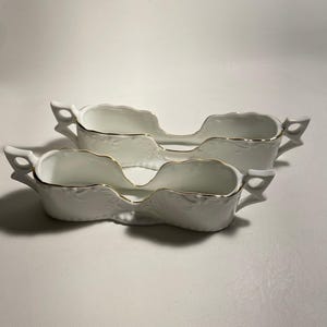 May include: Two white porcelain spoon rests with gold trim. The rests are shaped like a pair of spoons, with a raised design on the surface.