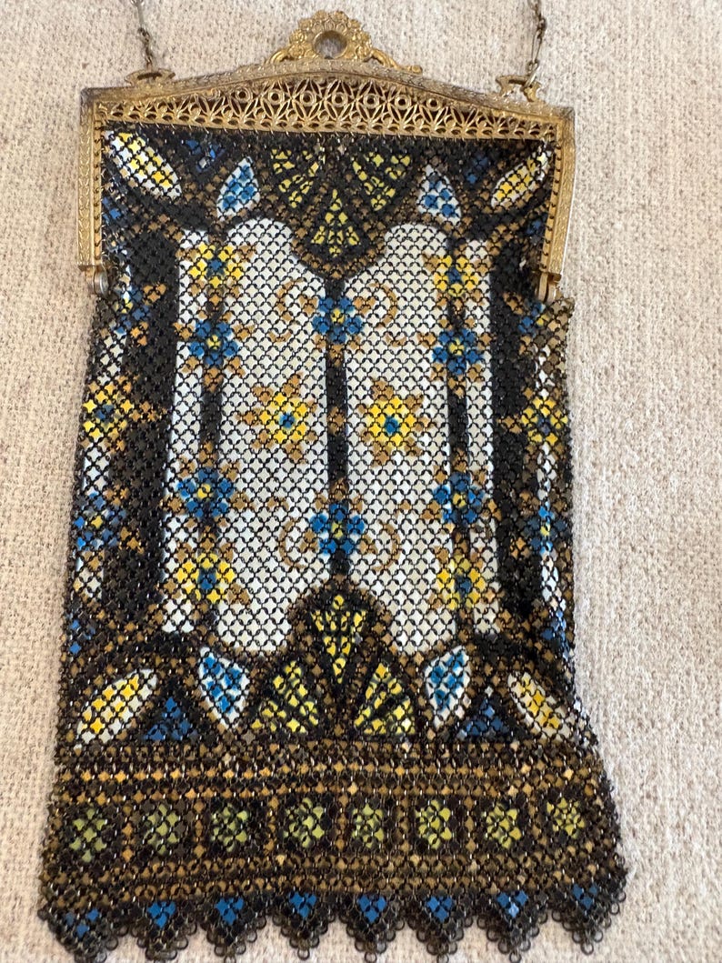 May include: An antique beaded handbag with a gold-tone frame and chain. The bag features a black mesh background with a geometric pattern in white, yellow, and blue. The bottom edge has a scalloped design.