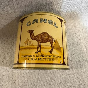 May include: Vintage Camel cigarette tin with a beige background. The tin features a brown camel, pyramids, and palm trees. The text "CAMEL" is at the top, and "TURKISH & DOMESTIC BLEND CIGARETTES" is at the bottom. The tin has a rectangular shape.