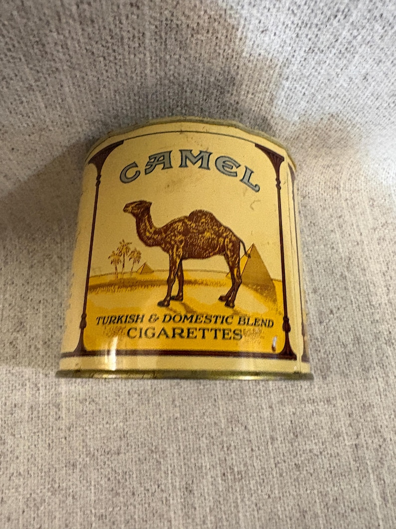 May include: Vintage Camel cigarette tin with a beige background. The tin features a brown camel, pyramids, and palm trees. The text "CAMEL" is at the top, and "TURKISH & DOMESTIC BLEND CIGARETTES" is at the bottom. The tin has a rectangular shape.
