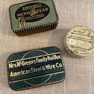 May include: Three antique metal tins. One is green and gold with "JOHN BULL MEND A TEAR" text. Another is a rectangular green and gold tin with "Mrs. McGregors Family Nail Box" text. A round tin with "PFLUEGER FISH HOOKS" text.