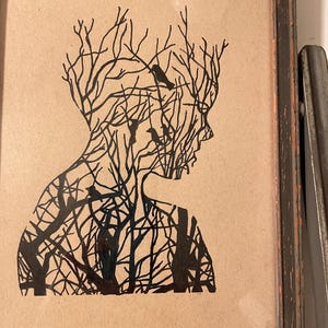 May include: A black and white silhouette of a person's head and shoulders, with a tree-like pattern inside. The silhouette is framed in a dark wood frame.