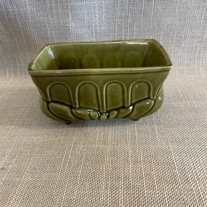 以下が含まれることがあります： 長方形のオリーブグリーンのセラミックプランター。ベースの周りに装飾的なアーチ型のデザインがあります。 プランターは光沢のある仕上がりで、小さな植物や多肉植物に適しています。