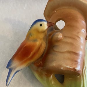 Puede incluir: Figura de pájaro de cerámica con cabeza azul, cuerpo amarillo y alas rojas, posada cerca de una casa para pájaros marrón. La casa para pájaros tiene una abertura redonda y está pintada en tonos marrones y beige. La base es verde.