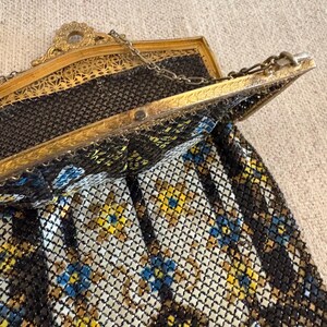 May include: An antique gold-toned metal mesh purse with a decorative frame. The purse features a chain handle and a pattern of black, yellow, and blue floral accents. The frame has intricate filigree details.