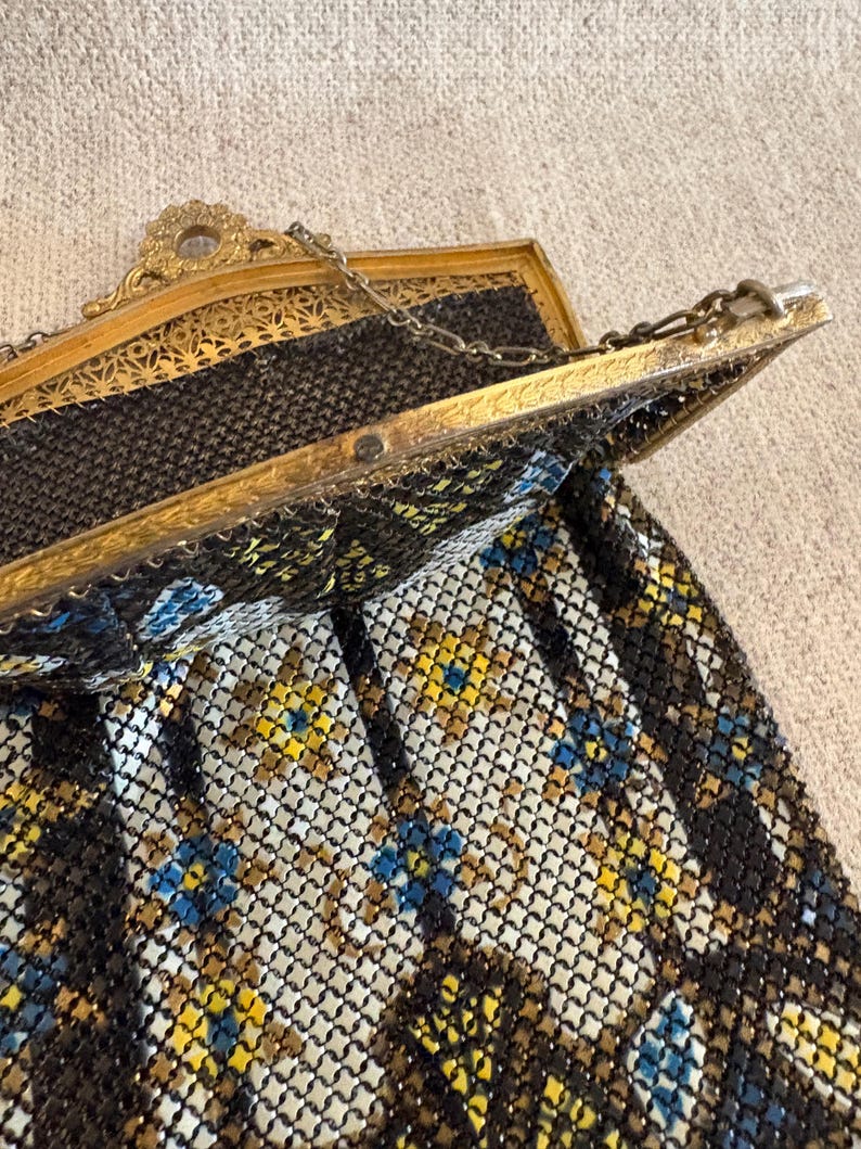 May include: An antique gold-toned metal mesh purse with a decorative frame. The purse features a chain handle and a pattern of black, yellow, and blue floral accents. The frame has intricate filigree details.