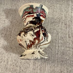 May include: A decorative ceramic vase with a unique, marbled design. The vase features a bulbous body and a flared top, with a colour palette of white, brown, red, and green. The base is a pedestal shape.