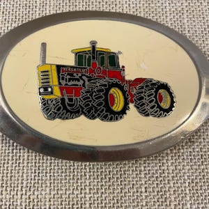 May include: Oval-shaped belt buckle with a tractor design. The tractor is red, yellow, and black, with the word "VERSATILE" on the side. The buckle has a silver-colored metal frame and a cream-colored background.