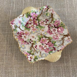 May include: A small, square ceramic dish with a scalloped edge. The dish features a floral pattern with pink blossoms and green leaves on a white background. The rim is trimmed with gold, and the dish has a light yellow base.