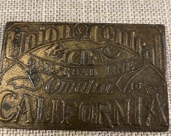 May include: An antique brass belt buckle with embossed text in a rectangular shape. The buckle reads "Union Central Pacific Railroad Line Omaha to California". It has a vintage, worn appearance.