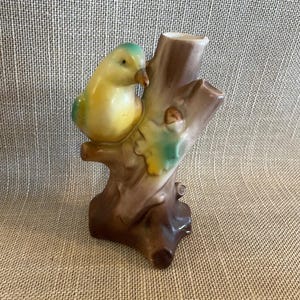 Puede incluir: Figura de pájaro de cerámica posada en un tocón de árbol marrón. El pájaro tiene colores amarillos y verdes, con un pico marrón. El tocón del árbol tiene una parte superior ahuecada y detalles verdes y marrones.