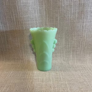 Op de afbeelding: Een lichtgroene glazen vaas met een geschulpte rand en decoratieve bladvormige patronen. De vaas heeft twee kleine handvatten en is ongeveer 15 cm hoog.