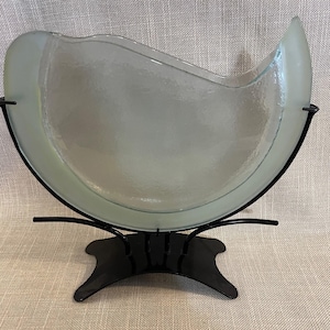 Könnte beinhalten: Eine Schale aus mattiertem Glas mit gewelltem Rand, getragen von einem schwarzen Metallständer. Die Schale hat einen hellgrünen Farbton und eine strukturierte Oberfläche. Der Ständer hat ein dekoratives, geschwungenes Design.